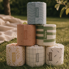 Six rolls of toilet paper stacked in a pyramid outdoors on green grass, each wrapped in a different patterned paper sleeve with a vertical 'Rollinsoft' label. The background shows a blurred park setting with trees and a blanket.